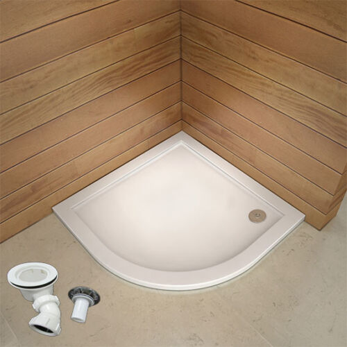 A white, slimline stone shower tray with a quadrant shape, shown installed in a bathroom setting with wood paneling in the background. A waste trap and piping are visible beside the tray.